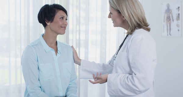 Ärztin berät lächelnde Patientin in der Praxis, eine Hand liegt auf der Schulter der Patientin.