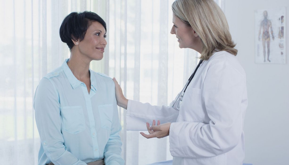Ärztin berät lächelnde Patientin in der Praxis, eine Hand liegt auf der Schulter der Patientin.