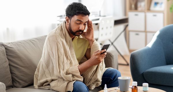 Ein Mann, der sich an die Schläfe fasst und ungesund aussieht, blickt eingehüllt in eine Decke auf sein Smartphone in seiner Hand. Vor ihm stehen auf einem kleinen Tisch eine Tasse sowie Medikamente.