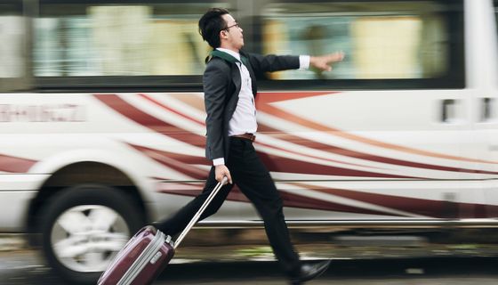 Junger Mann in Arbeitskleidung mit Ziehköfferchen rennt an einem Bus entlang.