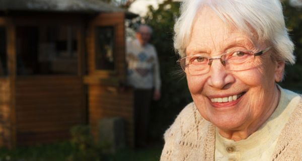 Porträt: Ältere Frau um die 80, beige Strickjacke, im Hintergrund Garten und Laube, lacht in die Kamera