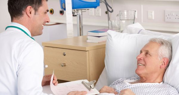 Älterer Patient im Krankenbett schaut lächelnd jüngeren Arzt an, der mit Krankenunterlagen an seinem Bett sitzt