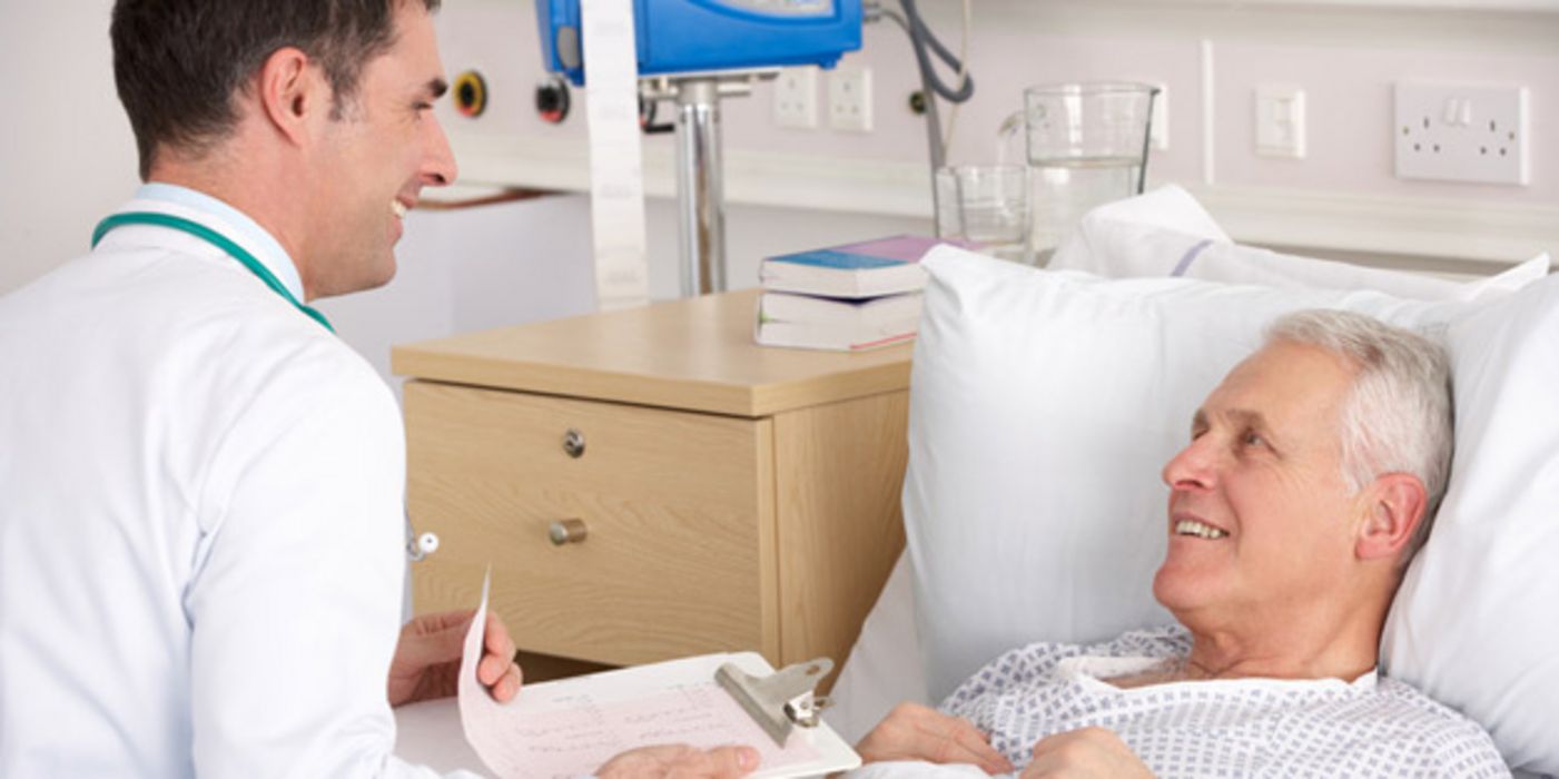 Älterer Patient im Krankenbett schaut lächelnd jüngeren Arzt an, der mit Krankenunterlagen an seinem Bett sitzt