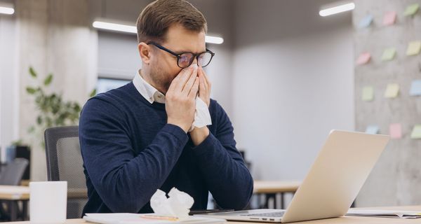 Junger Mann putzt sich im Büro die Nase. Vor ihm liegt bereits ein benutztes Taschentuch.