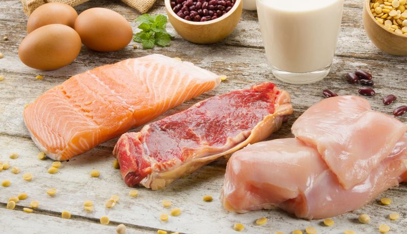 Auf einer Holzunterlage liegen drei Eier, ein Stück Lachs, Rindfleisch und Hühnerfleisch.