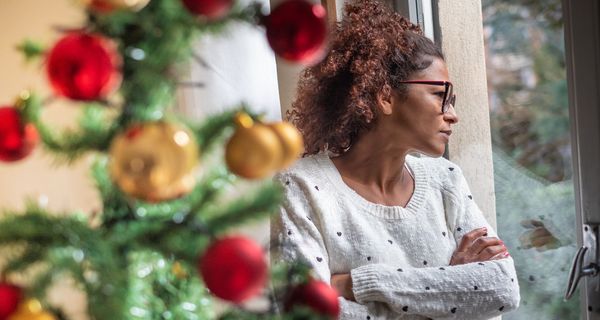 Frau in weihnachtlich dekoriertem Zimmer schaut nachdenklich aus Fenster