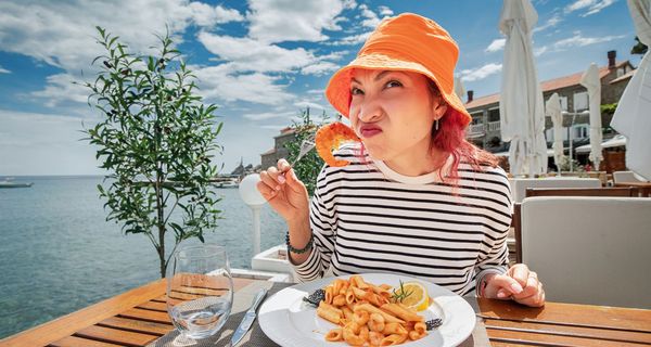 Frau mit orangem Hut isst Garnelen und rümpft skeptisch die Nase.