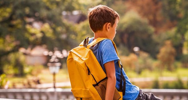 Junge mit Schulranzen, sitzt allein auf einer Mauer, Rückenansicht.