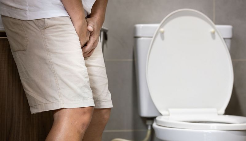 Jemand in Shorts hält sich den Unterleib, im Hintergrund eine Toilette.