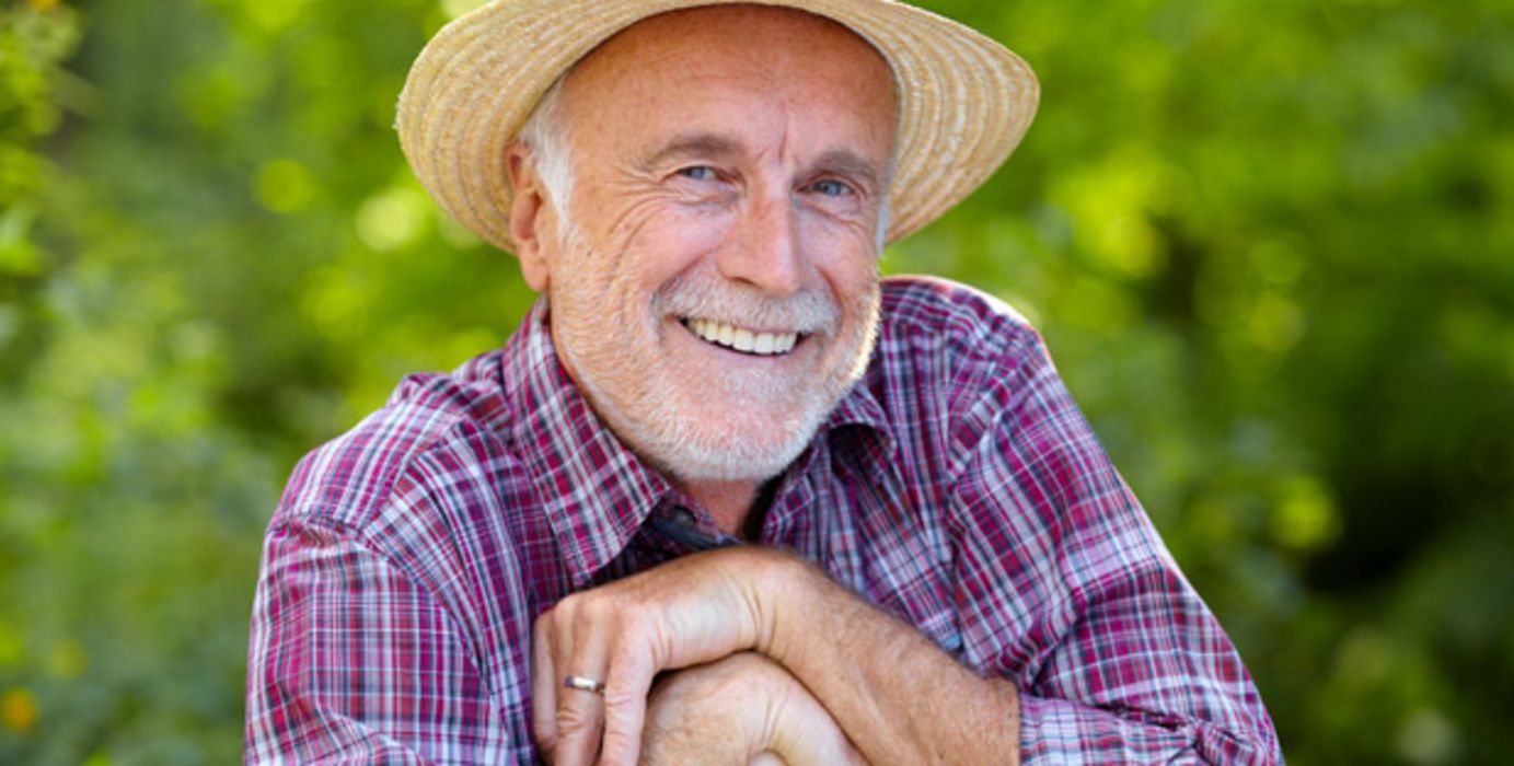 Älterer Mann, Karohemd, Strohhut, im Garten auf eine Schaufel gestützt lacht in die Kamera