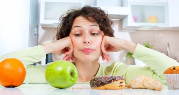 Junge Frau, stützt den Kopf auf die Hände, vor einer Orange, einem Apfel, einem Donut und einem Croissant.