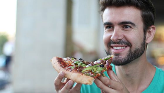 Foto Gesicht und Schulterbereich, junger dunkelhaariger, bärtiger Mann hält sich lächelnd ein großes Stück Pizza vor den Mund.