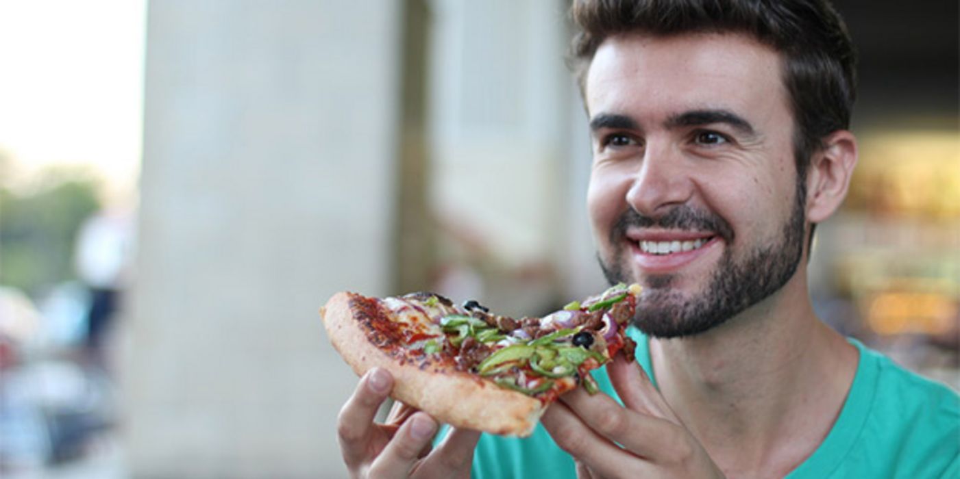 Foto Gesicht und Schulterbereich, junger dunkelhaariger, bärtiger Mann hält sich lächelnd ein großes Stück Pizza vor den Mund.