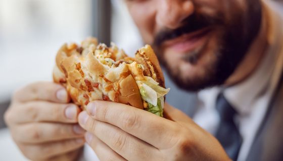 Mann hält einen Fast-Food-Burger, von dem er bereits abgebissen hat, in den Händen.