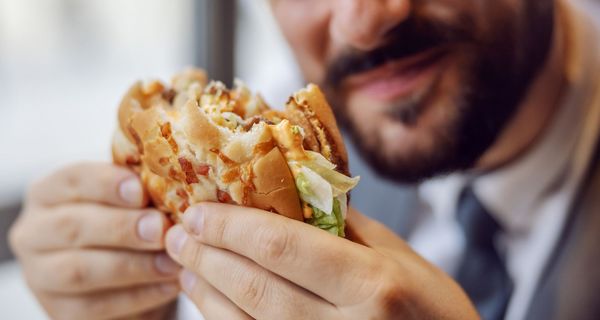 Mann hält einen Fast-Food-Burger, von dem er bereits abgebissen hat, in den Händen.