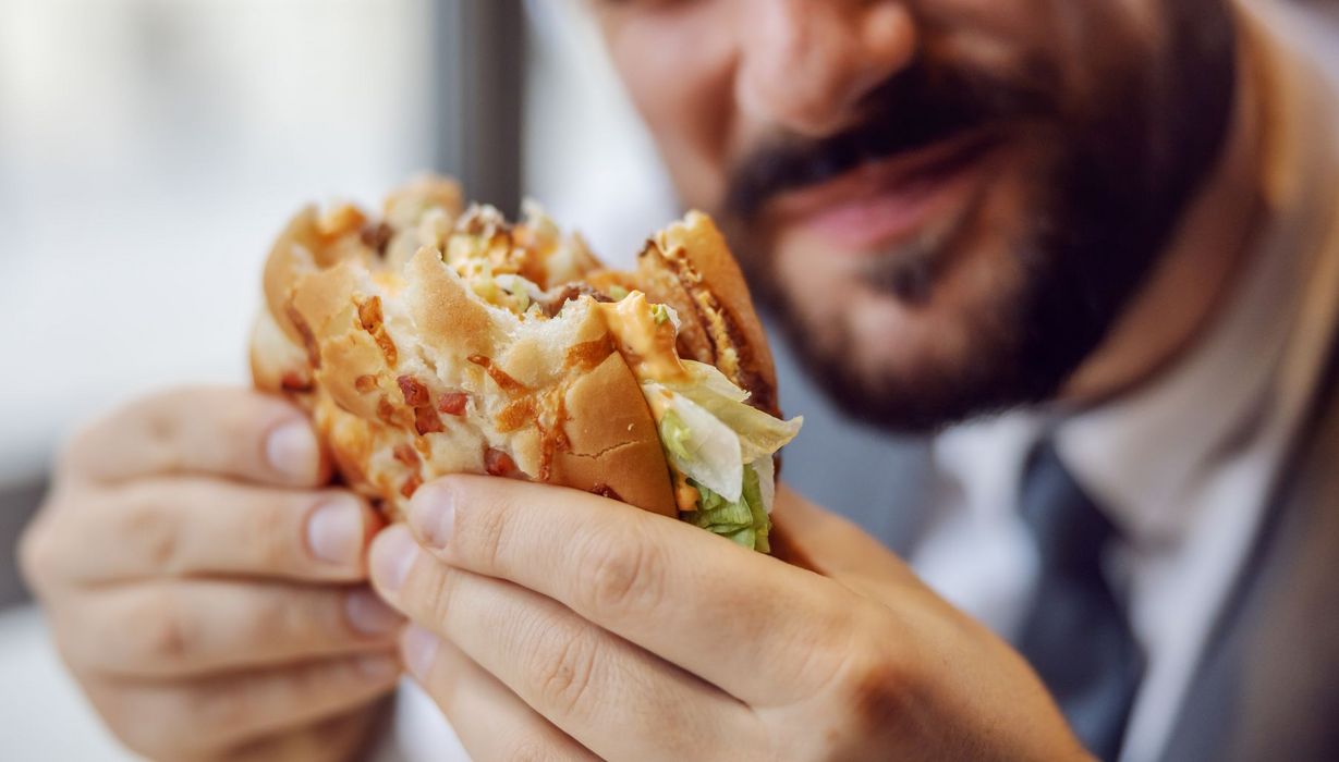 Mann hält einen Fast-Food-Burger, von dem er bereits abgebissen hat, in den Händen.