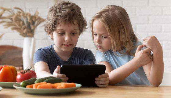 Ein Junge sitzt mit einem Tablet in der Hand an einem Tisch. Ein Mädchen sitzt neben ihm und schaut wie er auf den Bildschirm. Vor ihnen stehen Teller mit Gemüse 