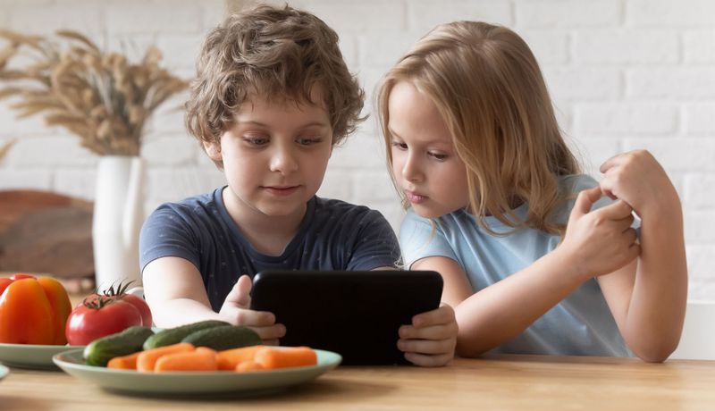 Ein Junge sitzt mit einem Tablet in der Hand an einem Tisch. Ein Mädchen sitzt neben ihm und schaut wie er auf den Bildschirm. Vor ihnen stehen Teller mit Gemüse