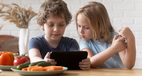 Ein Junge sitzt mit einem Tablet in der Hand an einem Tisch. Ein Mädchen sitzt neben ihm und schaut wie er auf den Bildschirm. Vor ihnen stehen Teller mit Gemüse