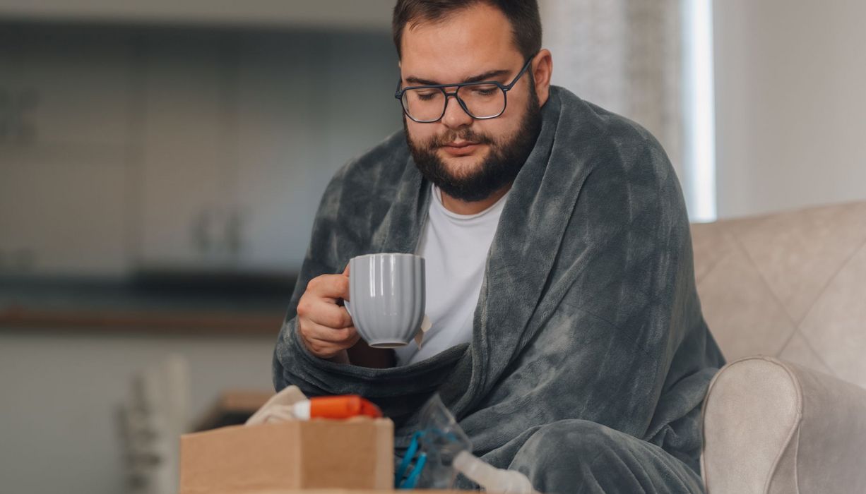 Mann mit Übergewicht, eingehüllt in eine Decke mit Tasse in der Hand, blickt auf einen Taschentuchspender und einen Inhalator.