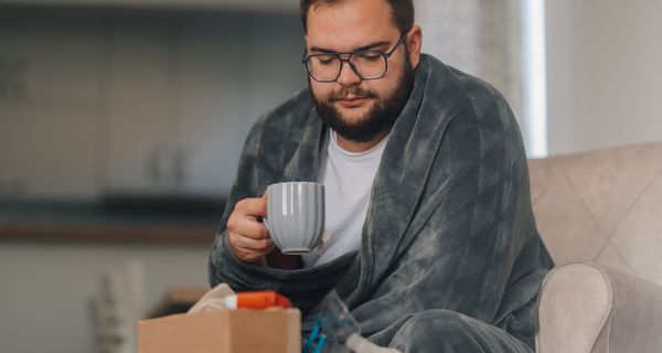 Mann mit Übergewicht, eingehüllt in eine Decke mit Tasse in der Hand, blickt auf einen Taschentuchspender und einen Inhalator.
