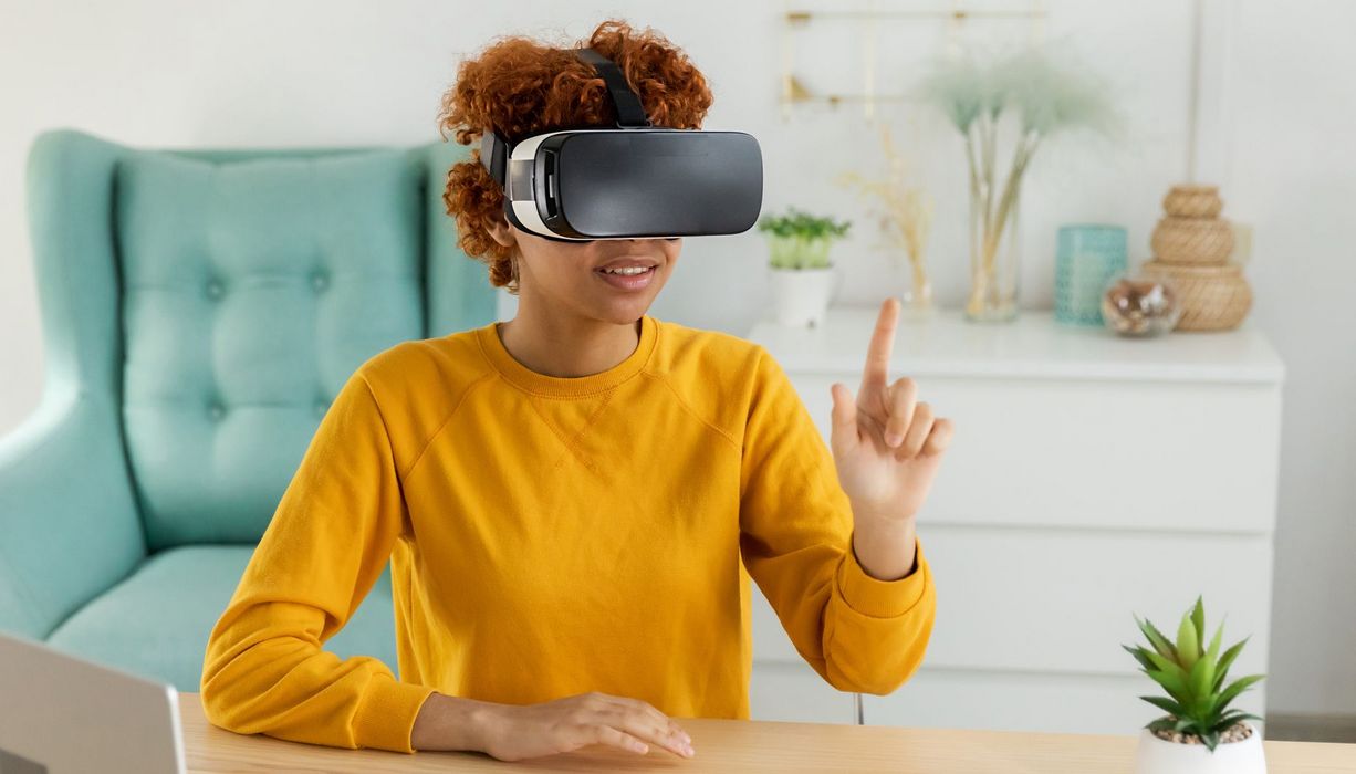 Eine junge Frau sitzt mit einer VR-Brille an einem Schreibtisch.