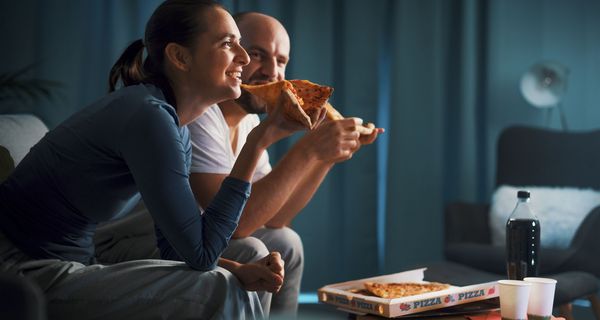 Paar, sitzt abends auf der Couch und isst Pizza.
