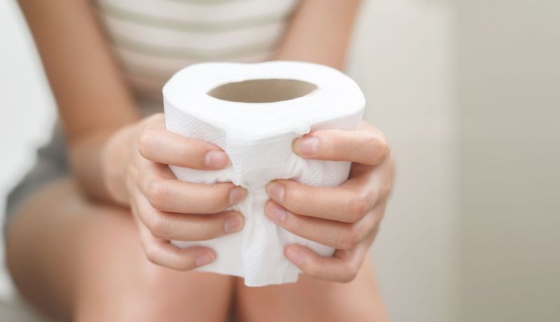 Frau sitzt auf der Toilette, hält eine Klopapierrolle fest. Ihr Gesicht ist nicht zu sehen. 