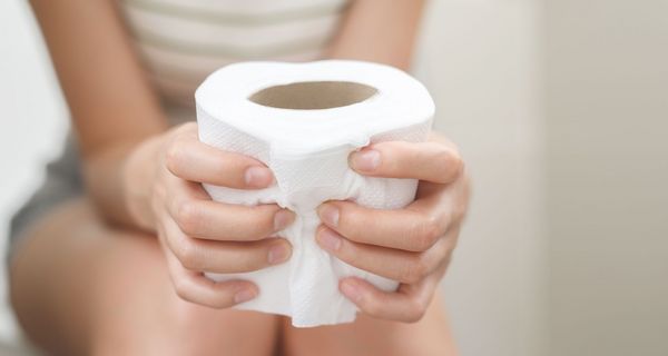 Frau sitzt auf der Toilette, hält eine Klopapierrolle fest. Ihr Gesicht ist nicht zu sehen. 