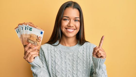 Strahlende Frau im Pullover hält ein Bündel 50-Euro-Scheine in der Hand.