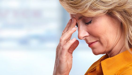 Rechter Bildrand Profil: Frau um die 60, blond, gelbe Bluse, Fingerspitzen der einen Hand fasst an schmerzende Stirn