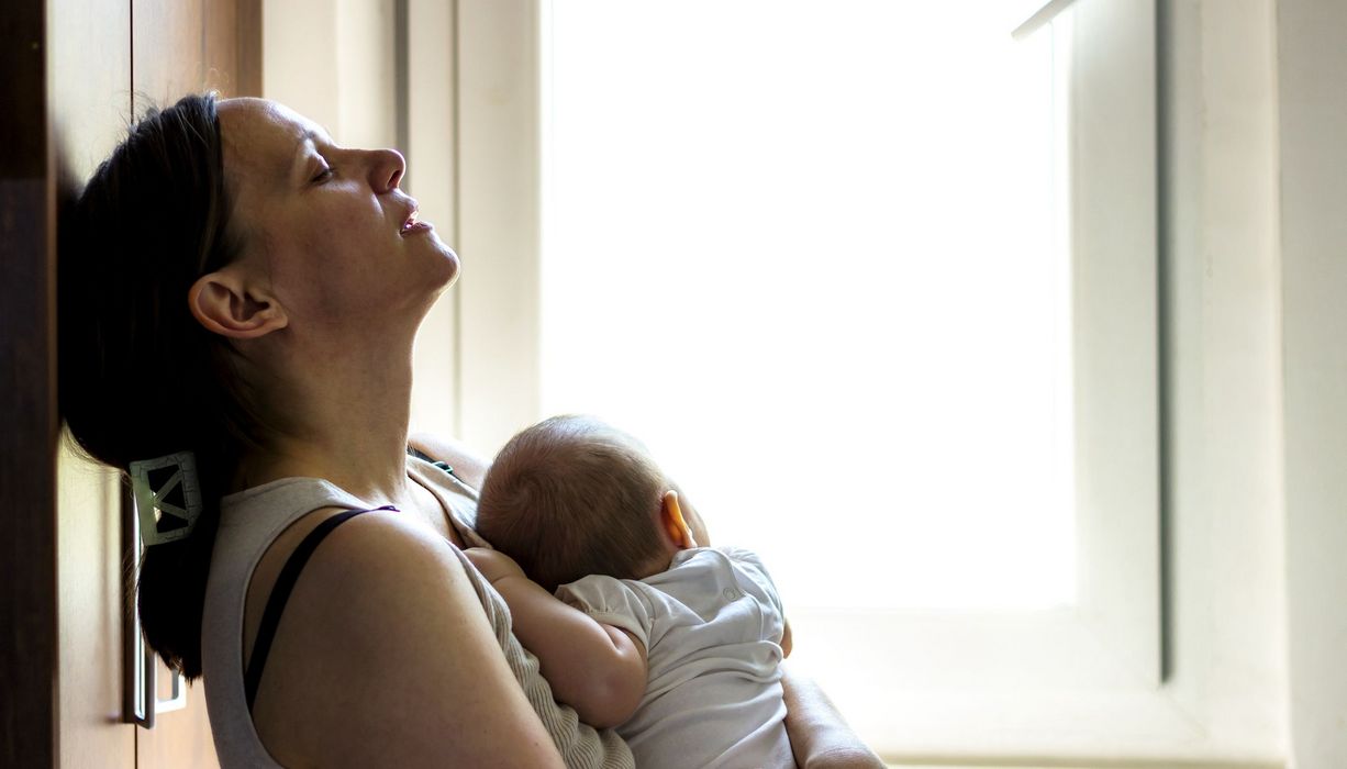 Mutter mit Baby im Arm, lehnt gestresst an der Wand.