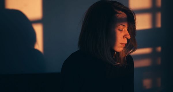 Frau, sitzt im Schatten eines Fensters und schaut nach unten.