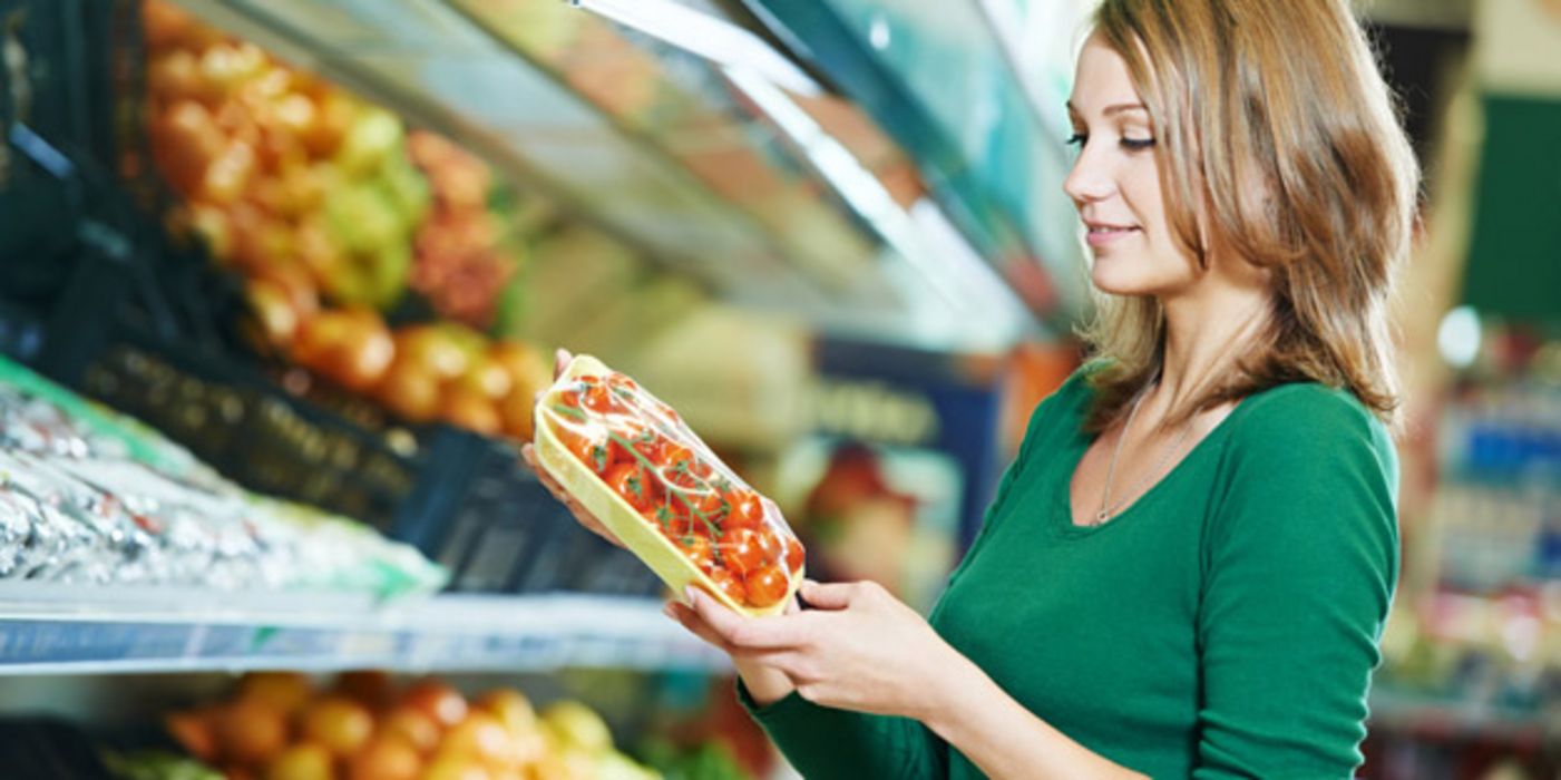 Junge Frau steht vor Supermarkt-Gemüseregal und hält eine Packung Tomaten in den Händen