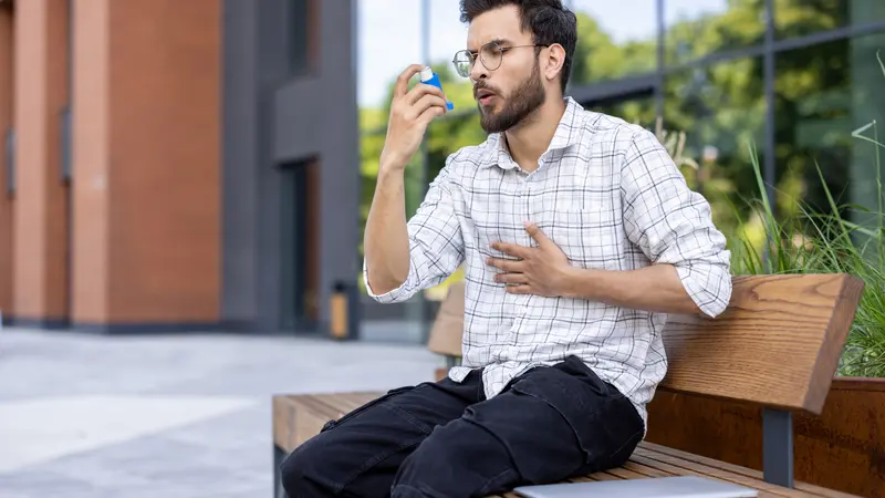 Ein junger Mann sitzt draußen, vor einem Gebäude auf einer Bank und benutzt einen Inhalator.