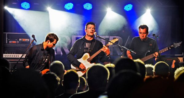 Drei Rockgitarristen auf der Bühne, Publikum im Vordergrund