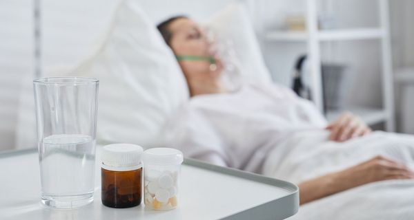 Frau im Krankenhausbett, im Vordergrund Tabletten und Wasser.