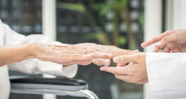 Eine Seniorin streckt einem Arzt im Kittel die Hände flach entgegen. Nur die Hände und Teile der Ärmel sind zu sehen. 