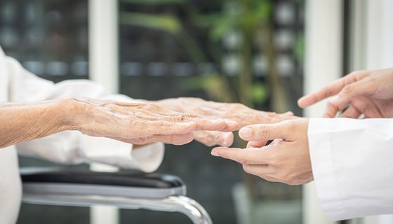 Eine Seniorin streckt einem Arzt im Kittel die Hände flach entgegen. Nur die Hände und Teile der Ärmel sind zu sehen. 