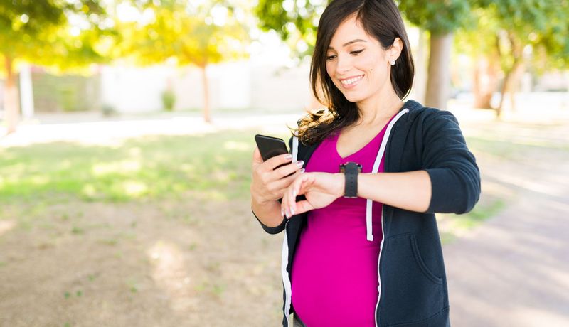 Schwangere Frau blickt auf ihre Smartwatch - Fitnesstracker - und ihr Smartphone. 