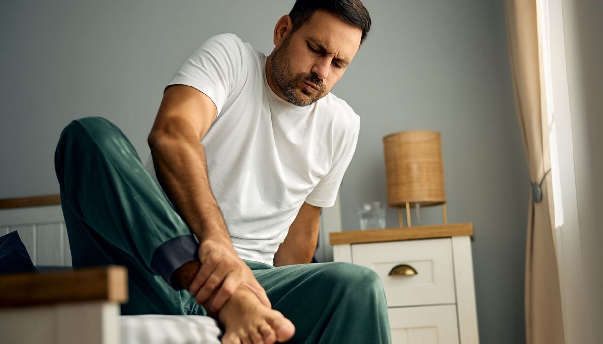 Mann steigt mit Schmerzen am Knöchelgelenk aus dem Bett.