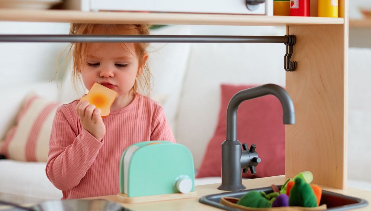 Kindergartenkind hält sich einen Spielzeugtoast an den Mund. Es steht hinter einer Spielzeugküche mit Toaster.
