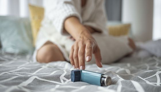 Eine FRau sitzt auf einem Bett und greift zu einem Asthma-Spray..