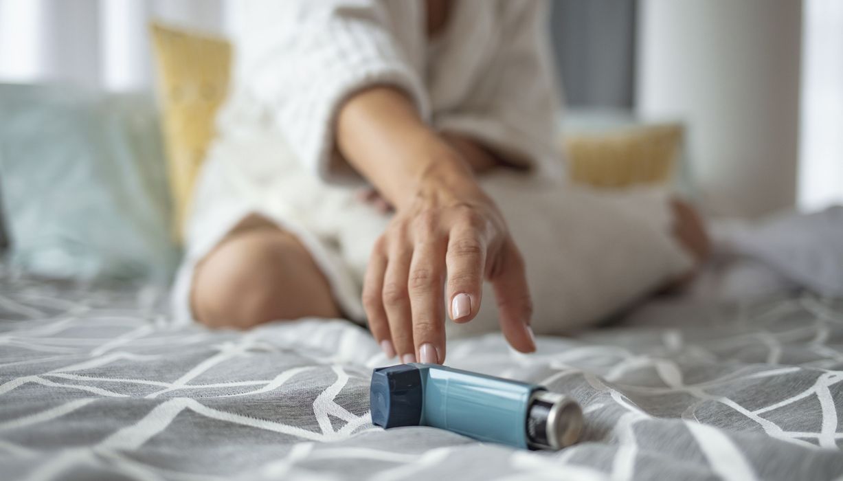 Eine FRau sitzt auf einem Bett und greift zu einem Asthma-Spray..