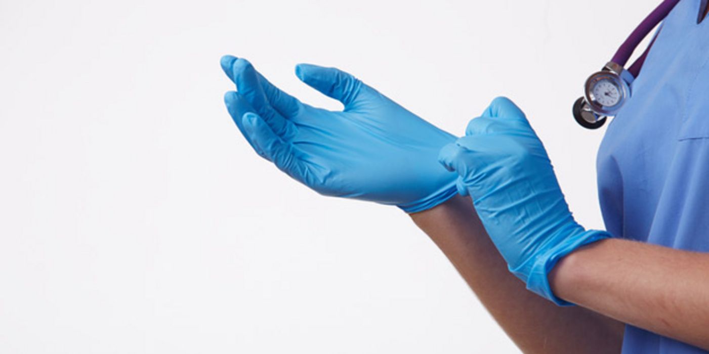 Blaue Einmalhandschuhe (Gummihandschuhe) werden von tw. zu sehender Person im blauen OP-Kittel mit Stethoskop um den Hals angezogen