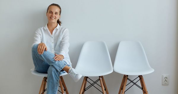 Eine junge Frau in weißem Hemd und Jeans sitzt auf einem Stuhl im Wartebereich. Sie lächelt.