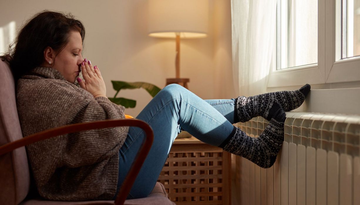 Frau sitzt auf einem Sessel vor der Heitzung, beide Füße in dicken Strümpfen wärmt sie am Heizkörper.