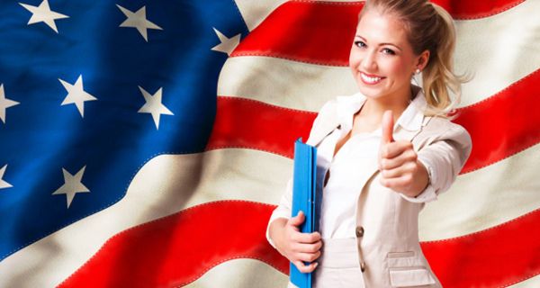 Junge, optimistische Amerikanerin vor einer US-Flagge reckt ihren Daumen nach oben