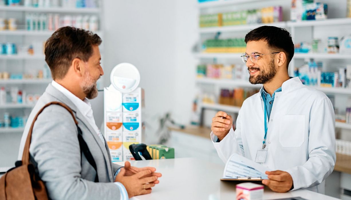 Ein Apotheker steht hinter einem HV-Tisch. Ein Kunde lässt sich von ihm zu Arzneimitteln beraten.