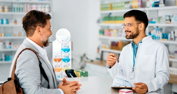 Ein Apotheker steht hinter einem HV-Tisch. Ein Kunde lässt sich von ihm zu Arzneimitteln beraten.